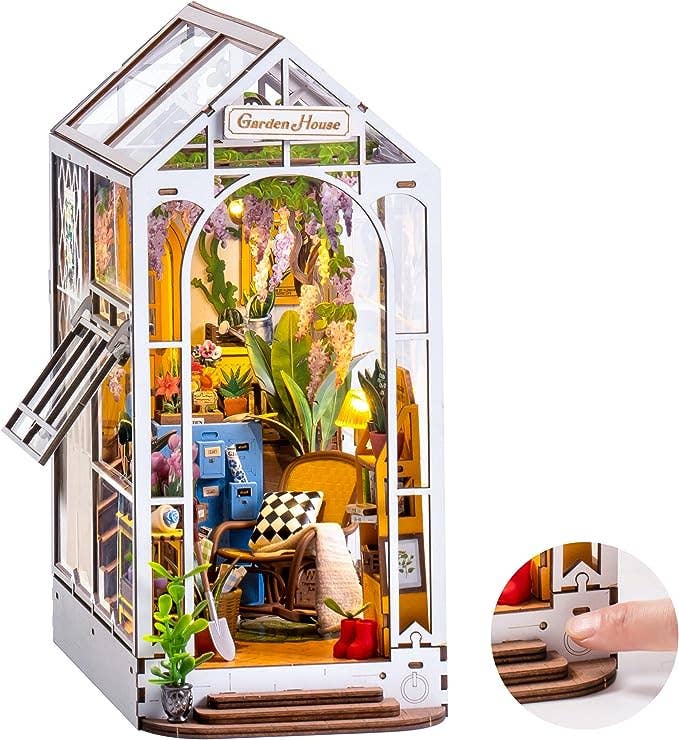 Miniature garden house model with open door, interior details, and a hand interacting with a small lever.