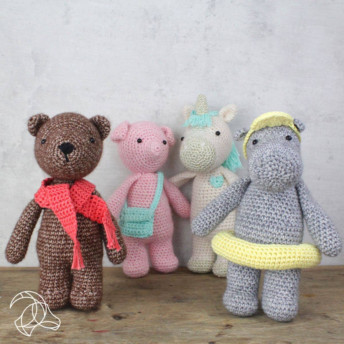 Four crocheted toys including a bear, pig, unicorn, and hippo on a wooden surface with a light gray background.