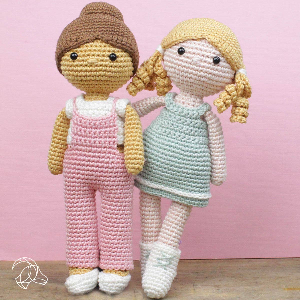 Two crochet dolls, one in pink overalls and the other in a light blue dress, against a pink background.