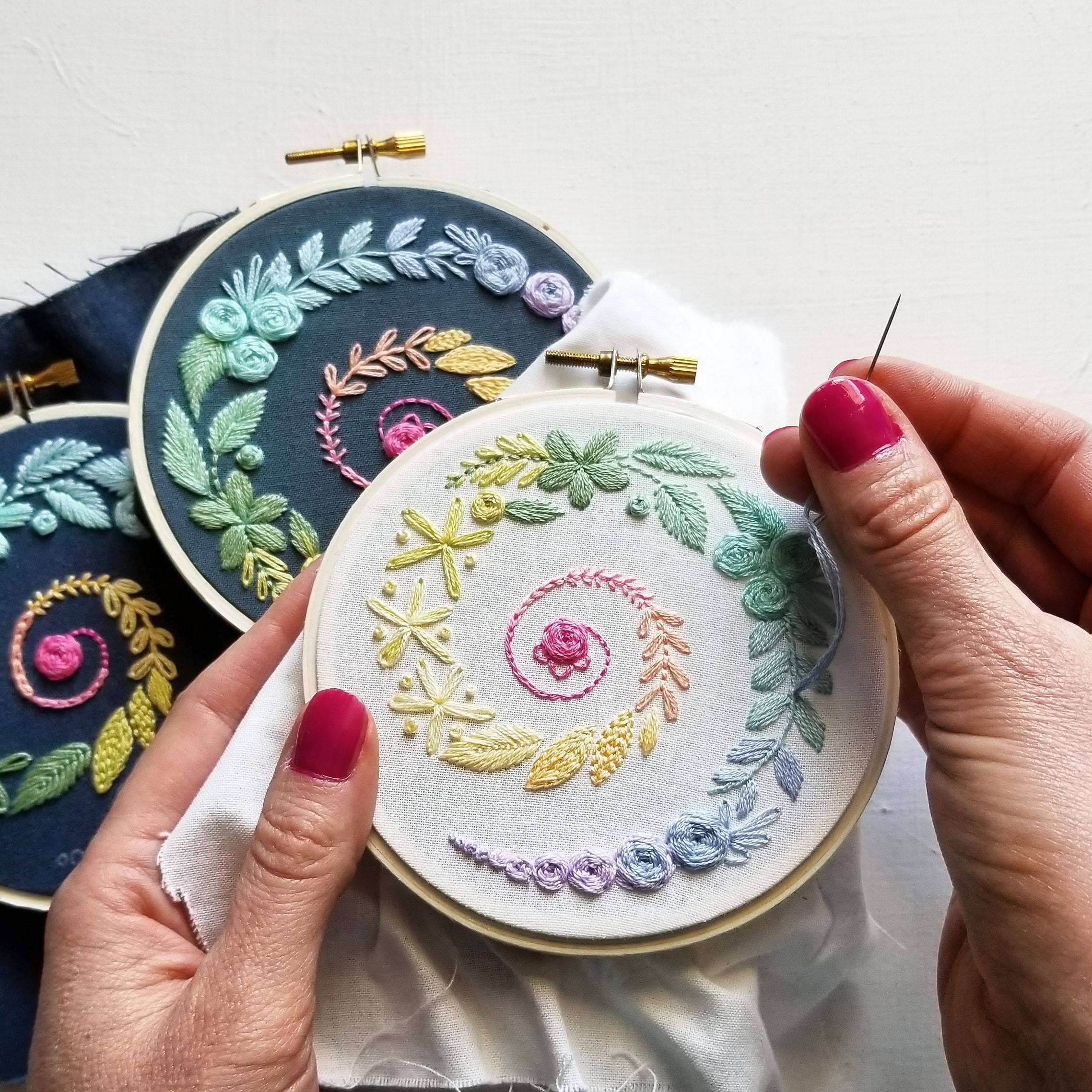 Embroidery hoop with colorful floral patterns held by hands with red nail polish.
