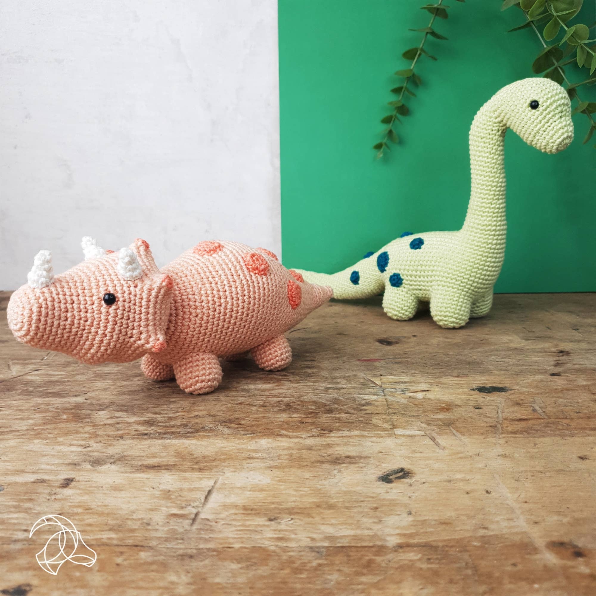 Two crochet dinosaurs, one pink and one green with blue spots, on a wooden surface with a green and white background.