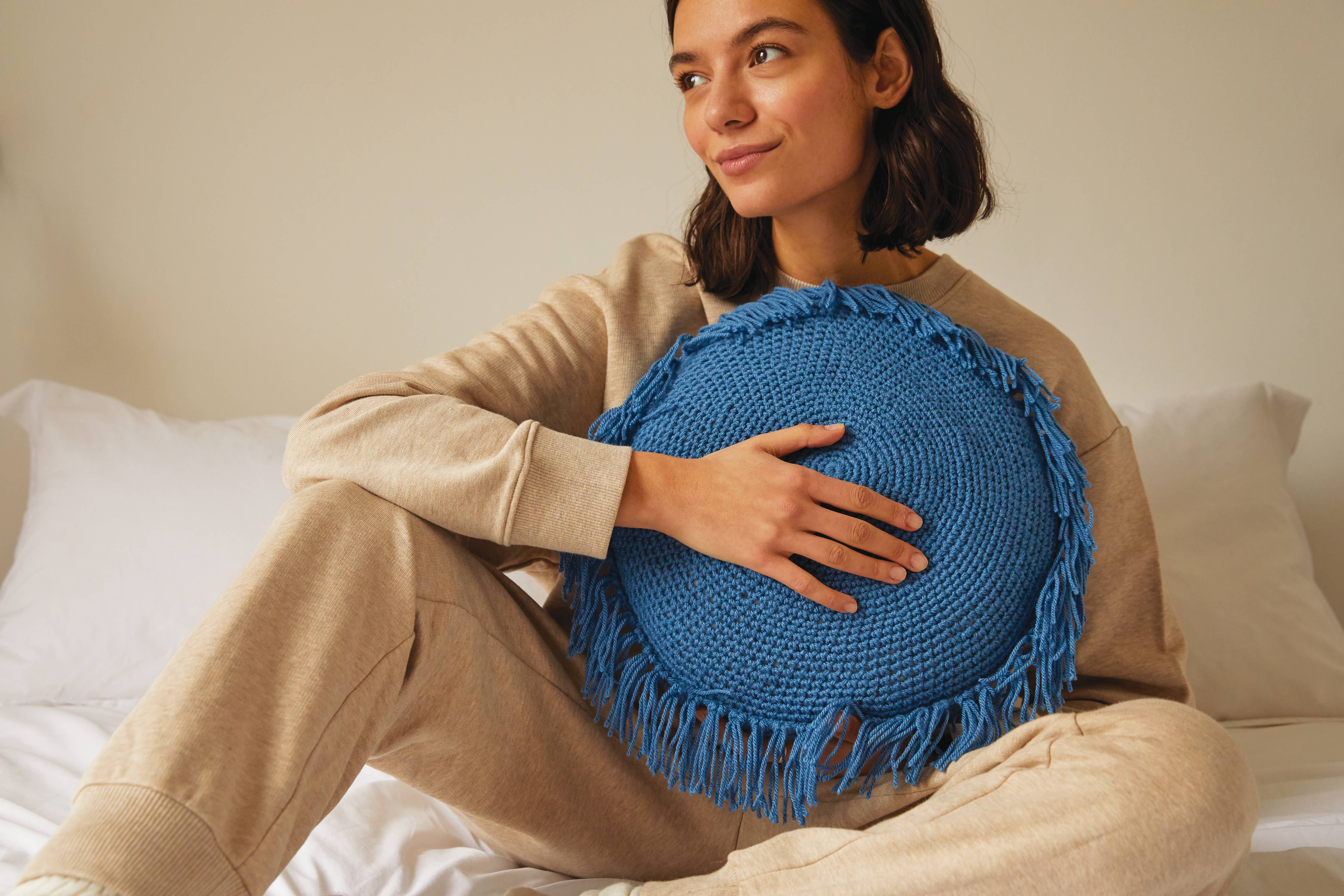 Woman holding a blue round pillow with tassels on a bed.