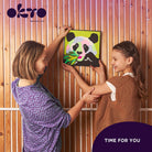 Two children holding a colorful painting of a panda against a wooden background with 'OKTO Sensory Art' branding.