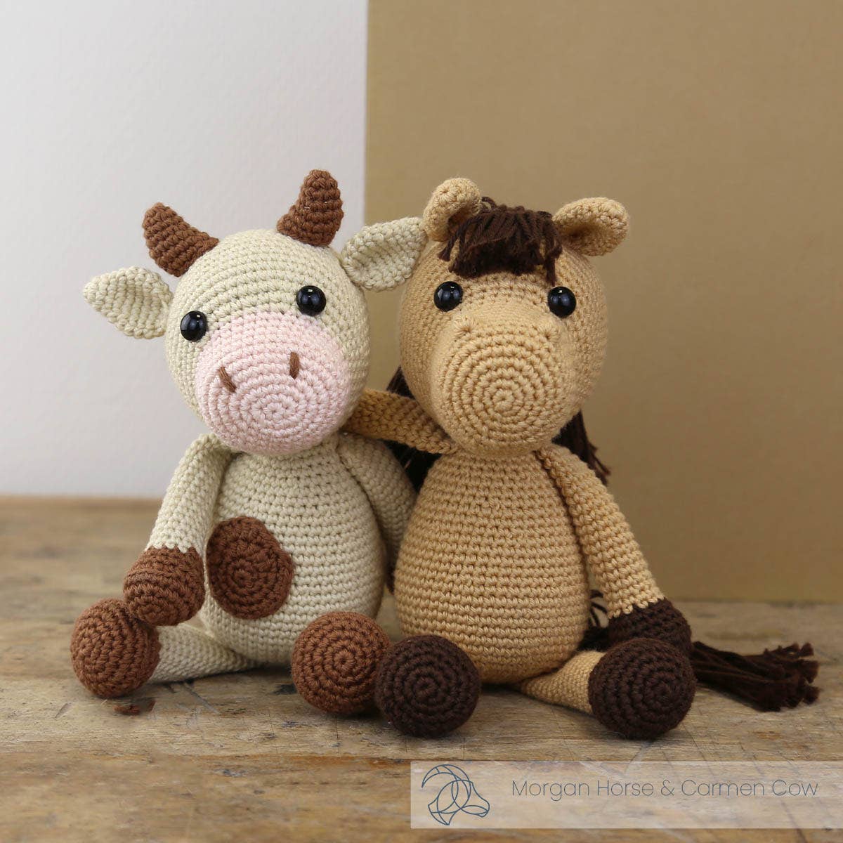 Crocheted horse and cow sitting together on a wooden surface with a neutral background.