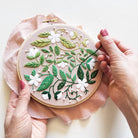 Embroidery hoop with green and white floral design held by hands on a light background