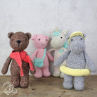 Four crocheted toys including a bear, pig, unicorn, and hippo on a wooden surface with a light gray background.