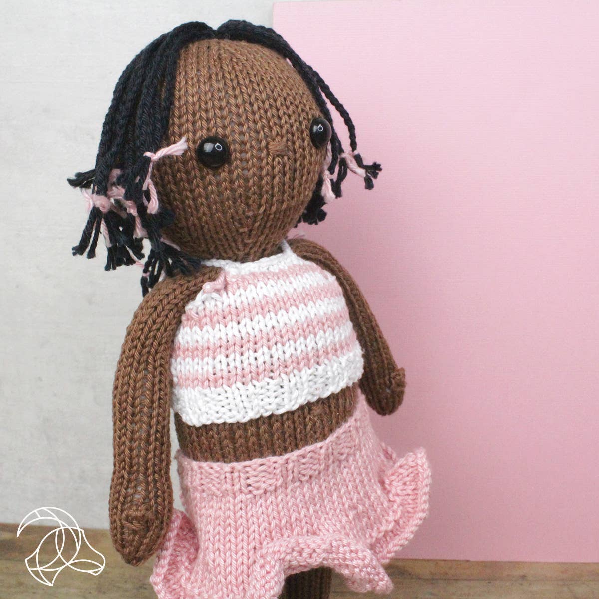 Knitted doll with braided hair wearing a pink and white striped shirt and pink skirt against a light pink background.