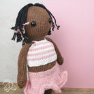 Knitted doll with braided hair wearing a pink and white striped shirt and pink skirt against a light pink background.