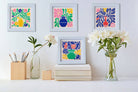 Colorful abstract art prints on a wall with flowers and books below.