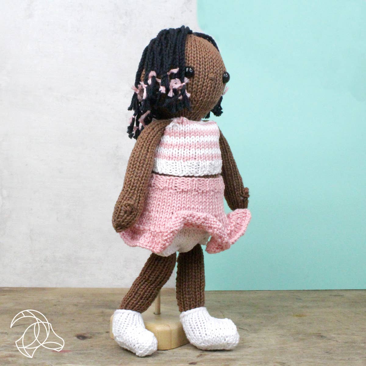 Knitted doll with braided hair wearing a pink cardigan, pink skirt, and white shoes on a wooden surface.