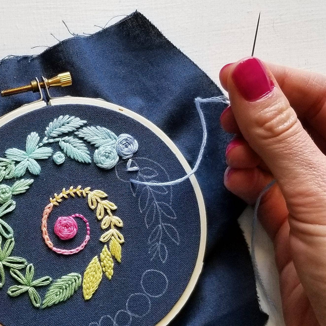 Embroidery hoop with floral design on blue fabric, hand holding a needle.