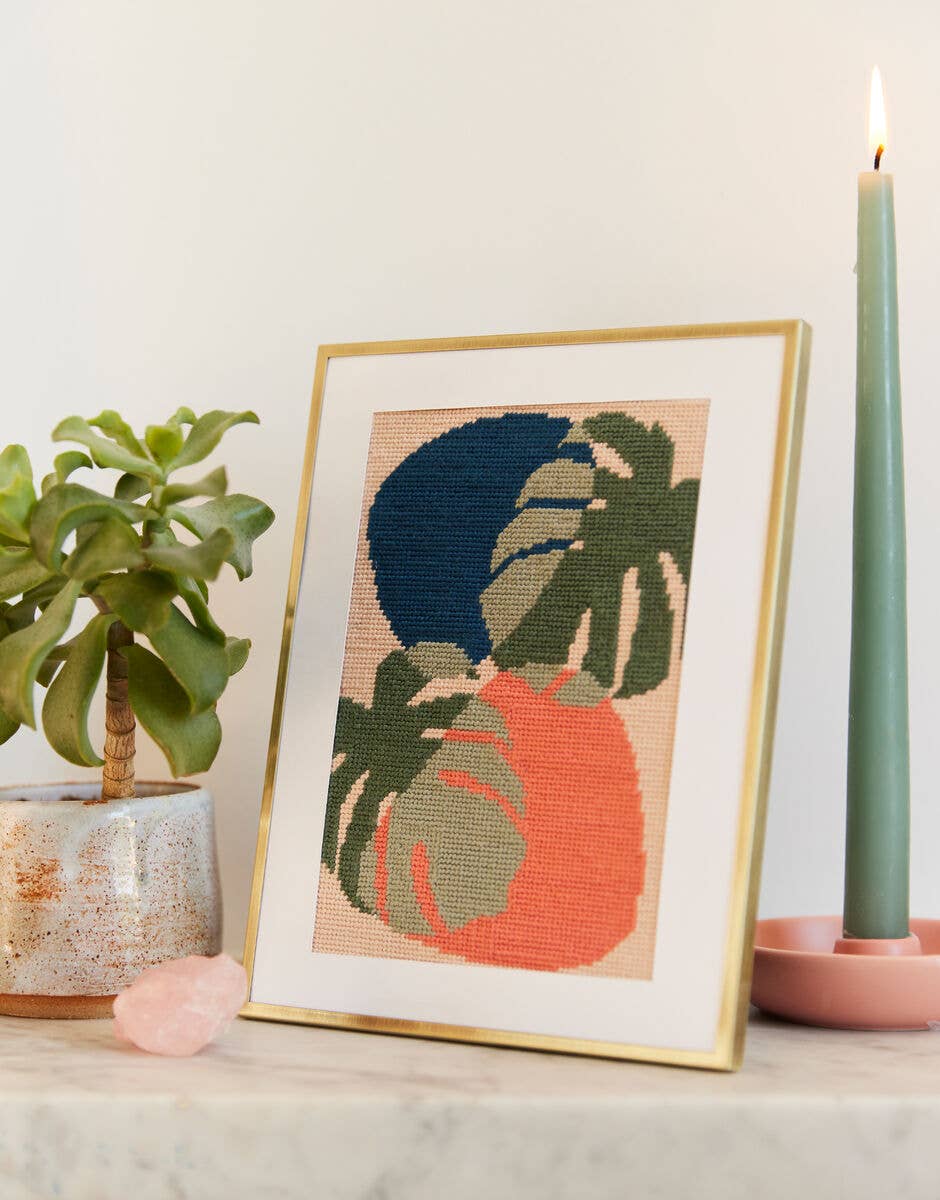 Framed cross-stitch art of tropical leaves on a table with a plant and candle.