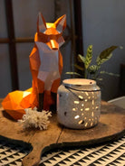 Geometric fox sculpture, white ceramic lantern, and coral on a wooden surface with a plant in the background.