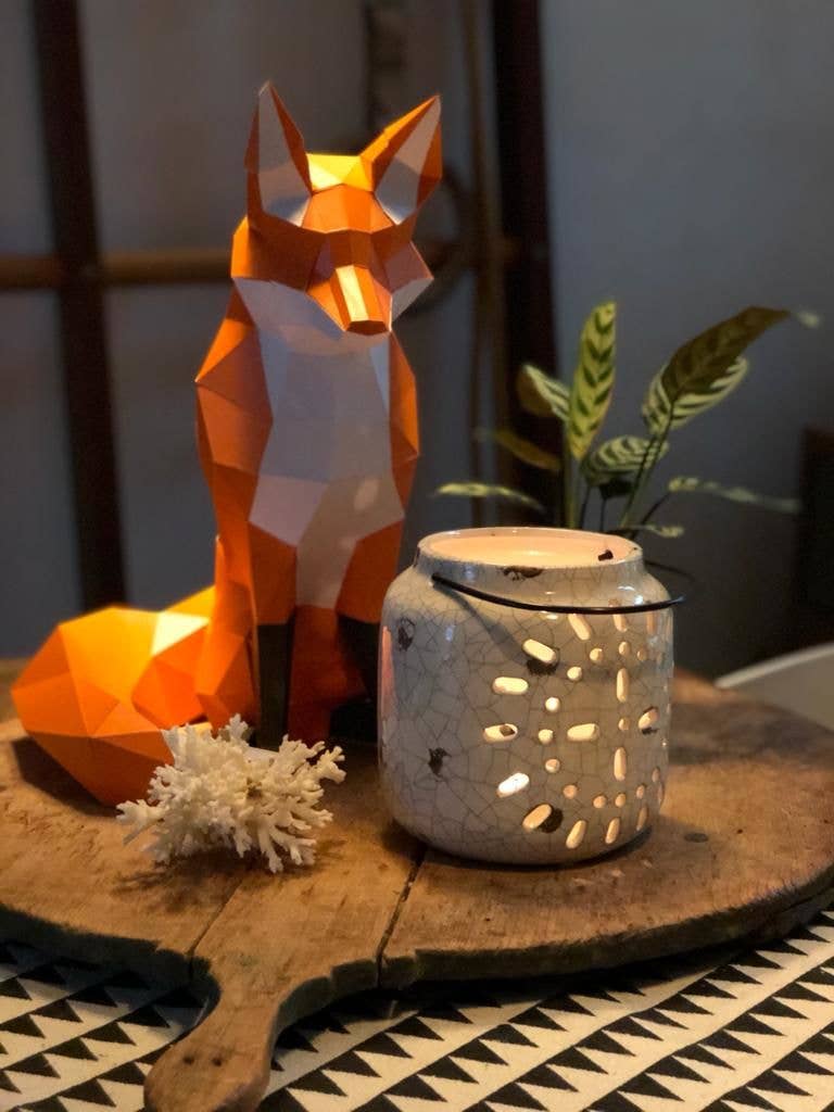 Geometric fox sculpture, white ceramic lantern, and coral on a wooden surface with a plant in the background.