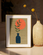 Framed needlepoint artwork of a blue vase with green leaves and an orange sun, next to a textured vase.