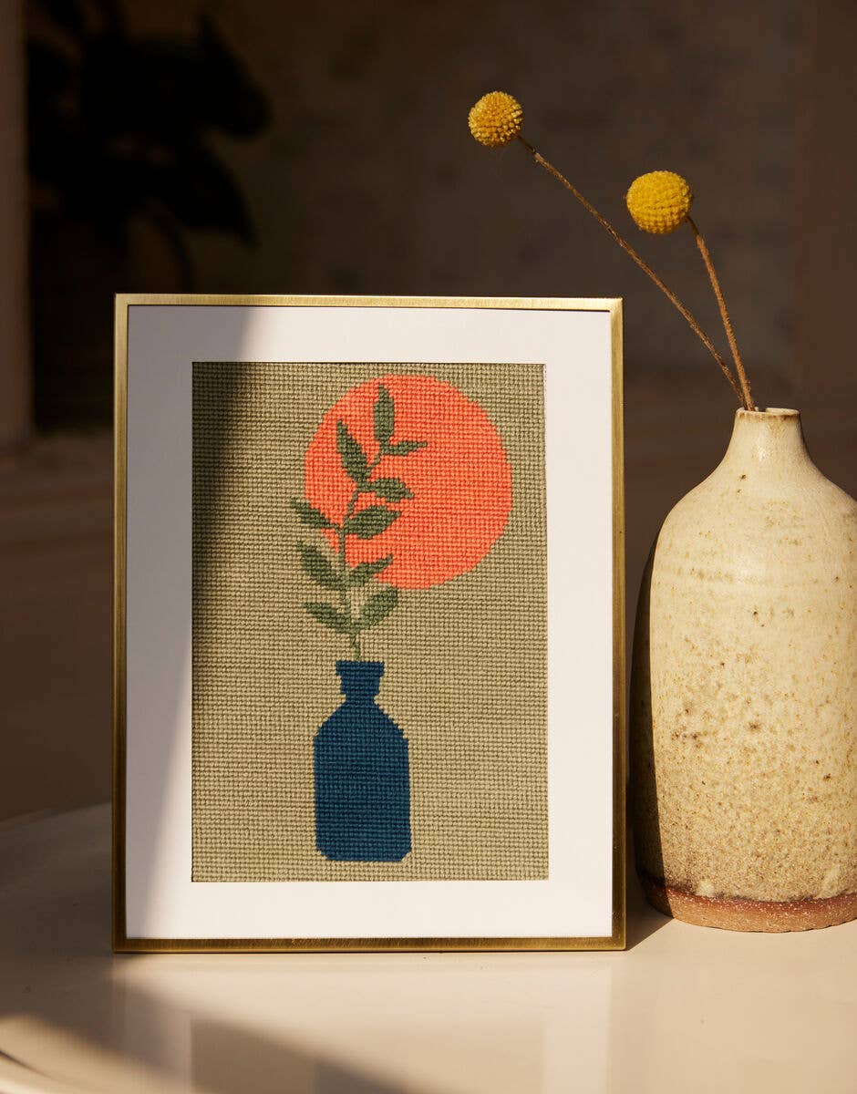Framed needlepoint artwork of a blue vase with green leaves and an orange sun, next to a textured vase.