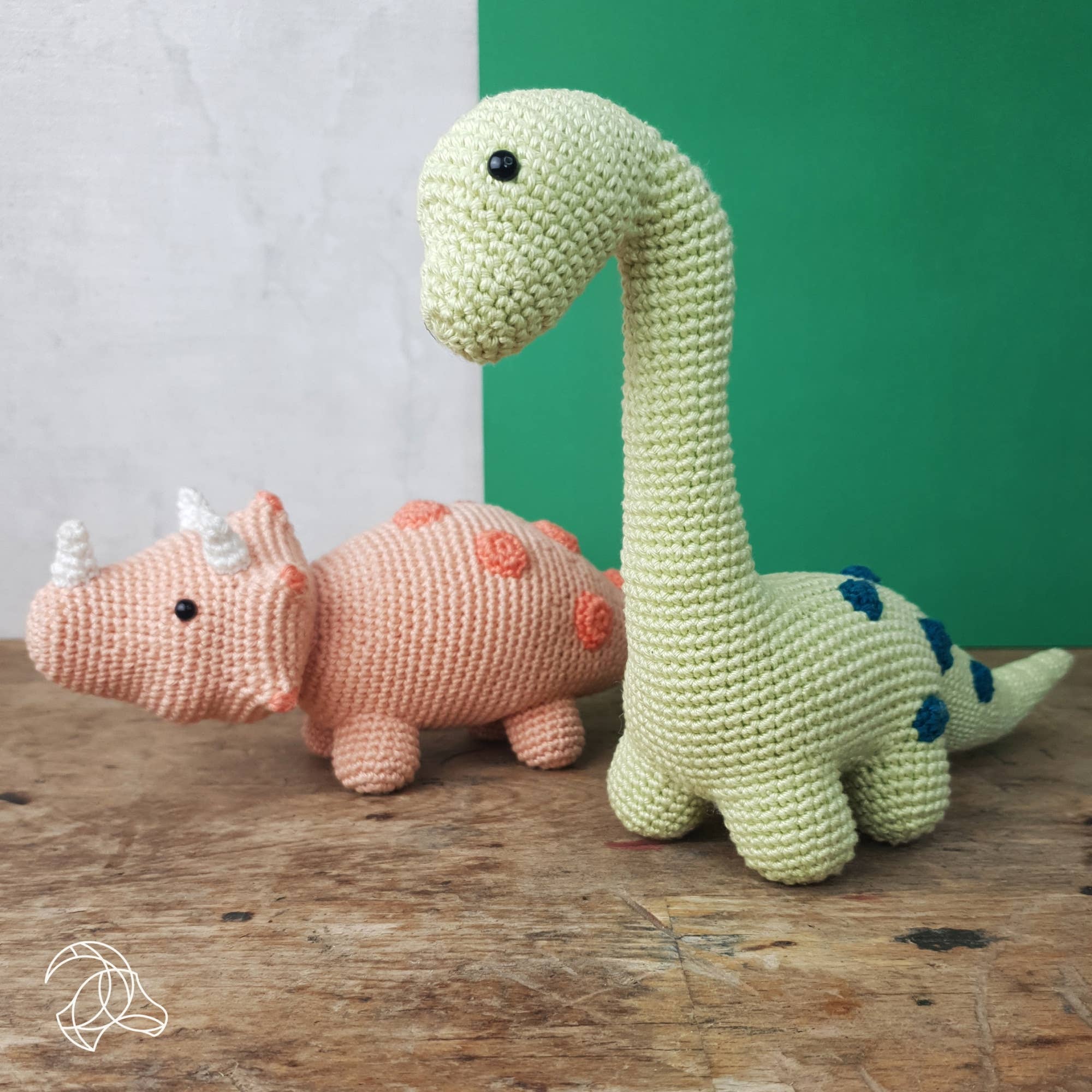 Two crochet dinosaurs, one green and one pink, on a wooden surface with a green wall in the background.