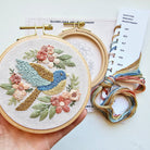 Embroidery hoop with colorful bird and floral design, held by a hand, with embroidery thread and pattern book in the background.