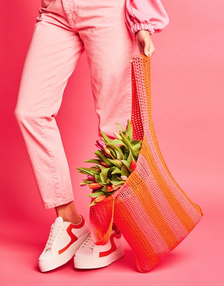 Person holding a colorful mesh bag with flowers against a pink background