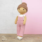 Crocheted doll with pink overalls and brown hair against a light pink background