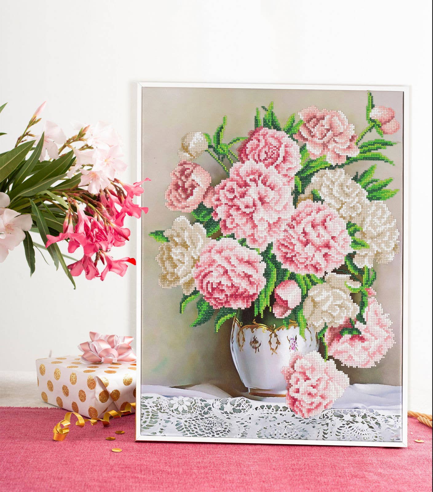 Framed needlepoint of pink and white flowers in a teacup on a pink surface with additional flowers and gift box.
