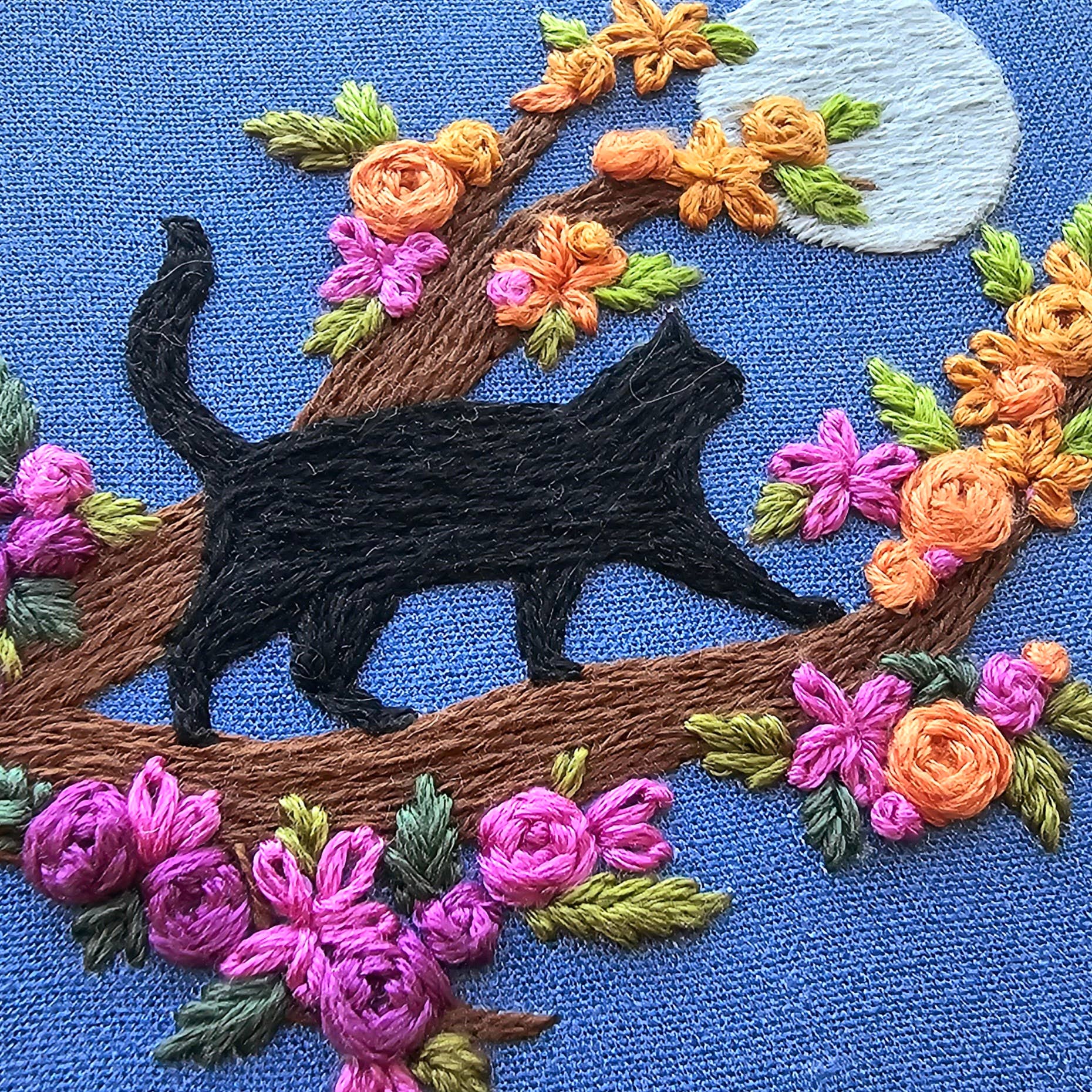 Embroidered design of a black cat among flowers on blue fabric