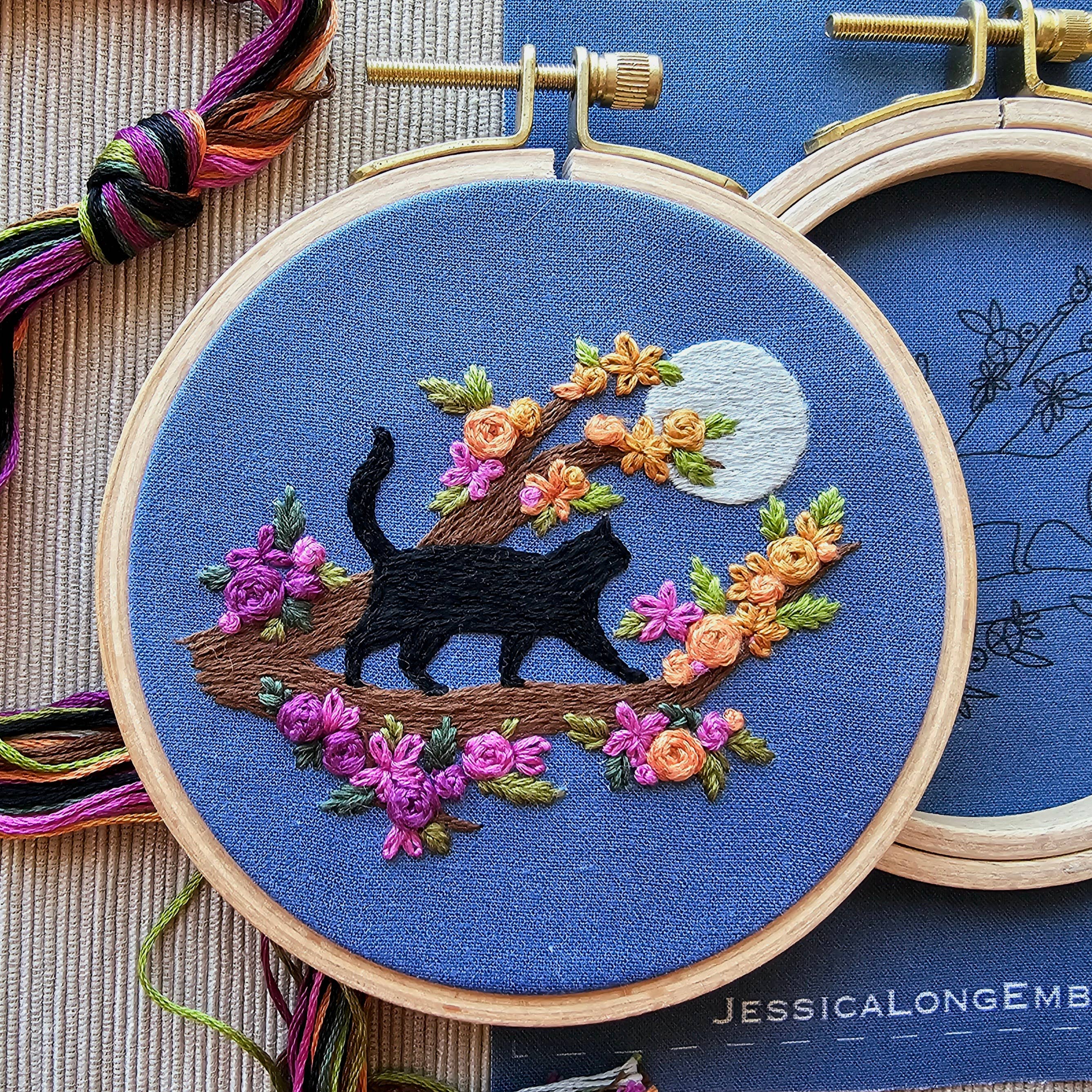 Embroidery hoop with a black cat design on blue fabric, surrounded by colorful flowers and leaves.