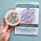 Embroidery hoop with bluebird design next to an embroidery kit packaging on a light blue background
