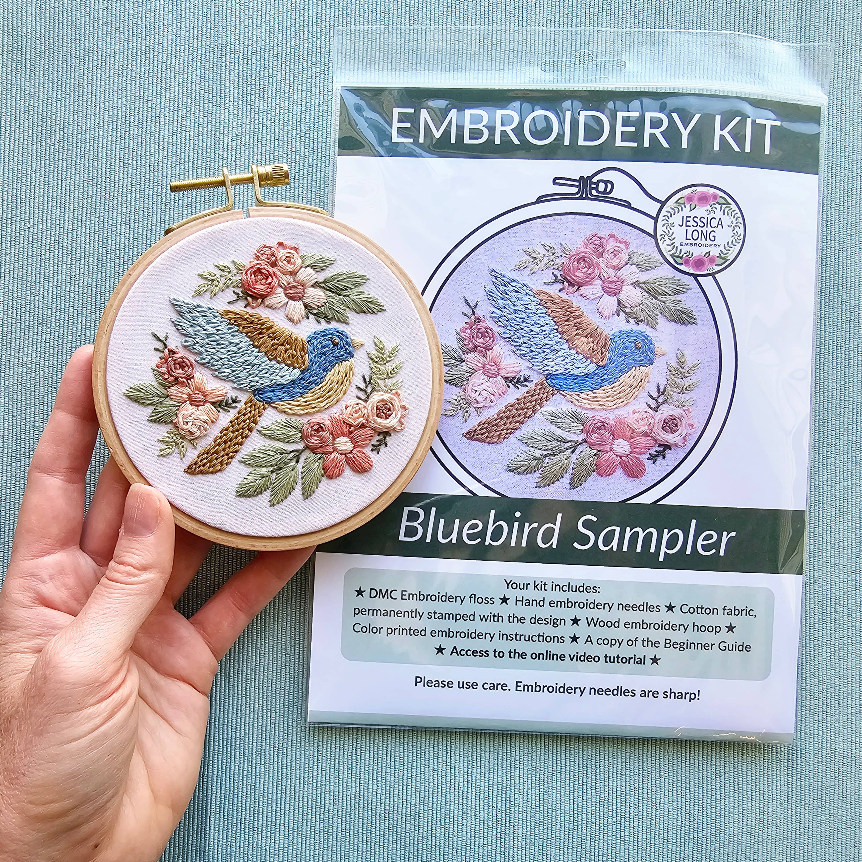 Embroidery hoop with bluebird design next to an embroidery kit packaging on a light blue background