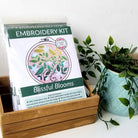 Embroidery kit with 'Blissful Blooms' design on a white surface with a plant.