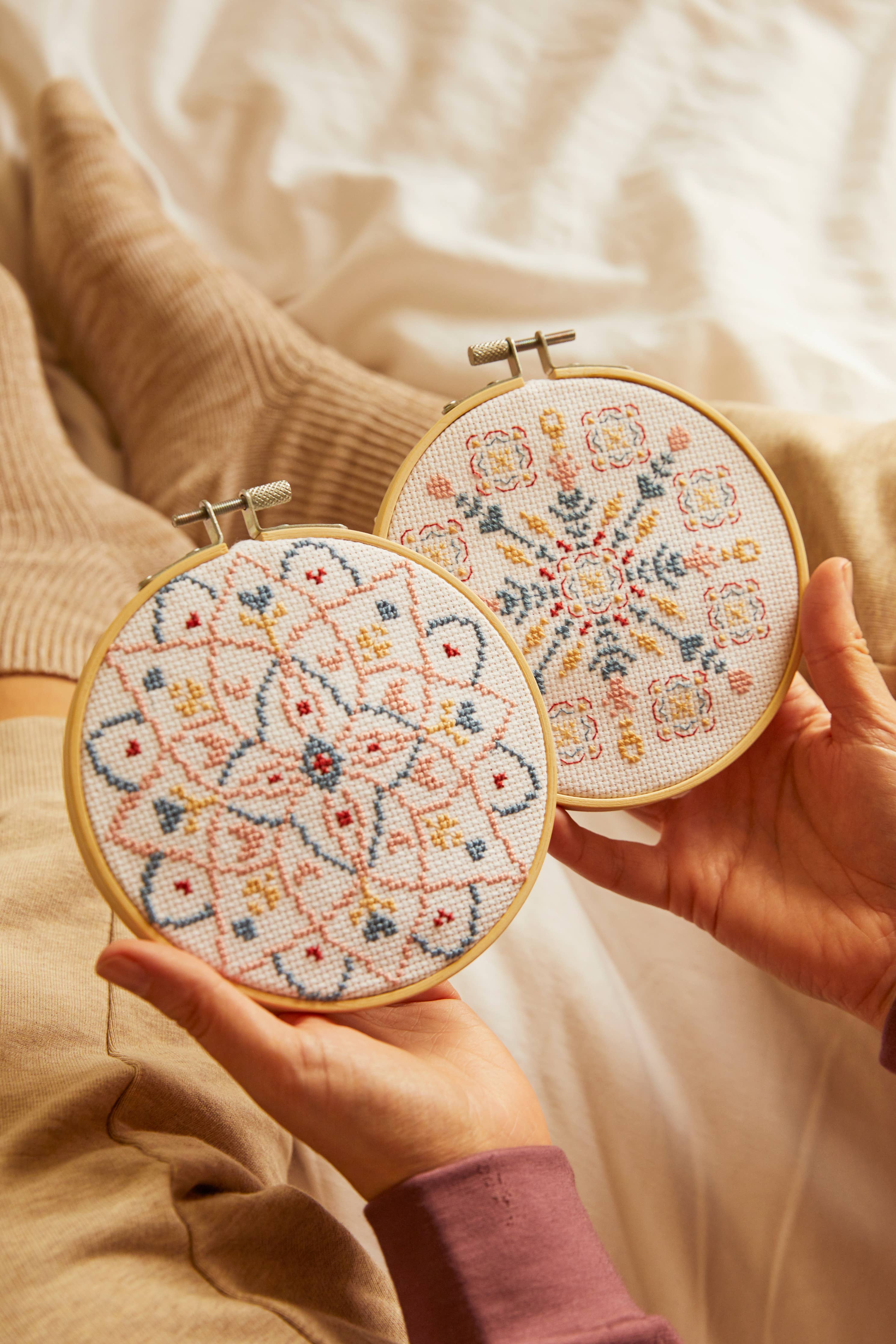Two hands holding embroidery hoops with floral patterns on a soft beige background