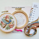 Embroidery hoops with floral and bird design, thread, and a pattern book on a white surface.