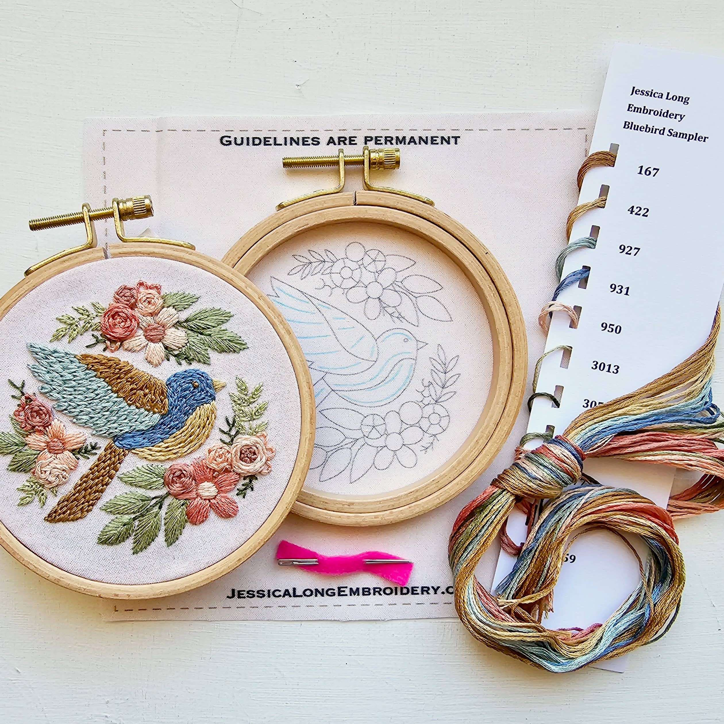 Embroidery hoops with floral and bird design, thread, and a pattern book on a white surface.