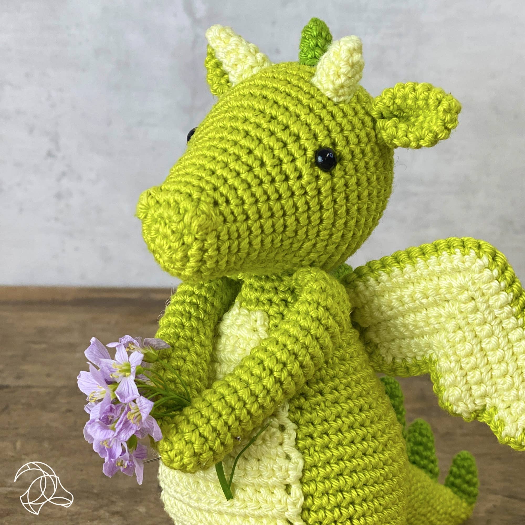 Green crochet dragon holding a purple flower on a wooden surface with a light gray background
