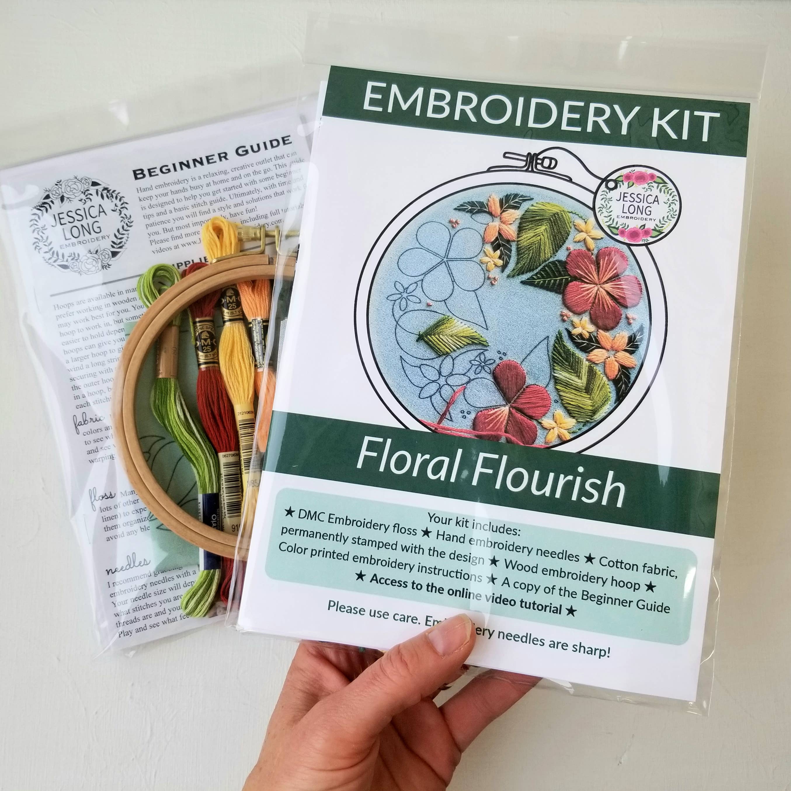 Embroidery kit with floral design held by a hand, including a beginner guide and sample hoop.