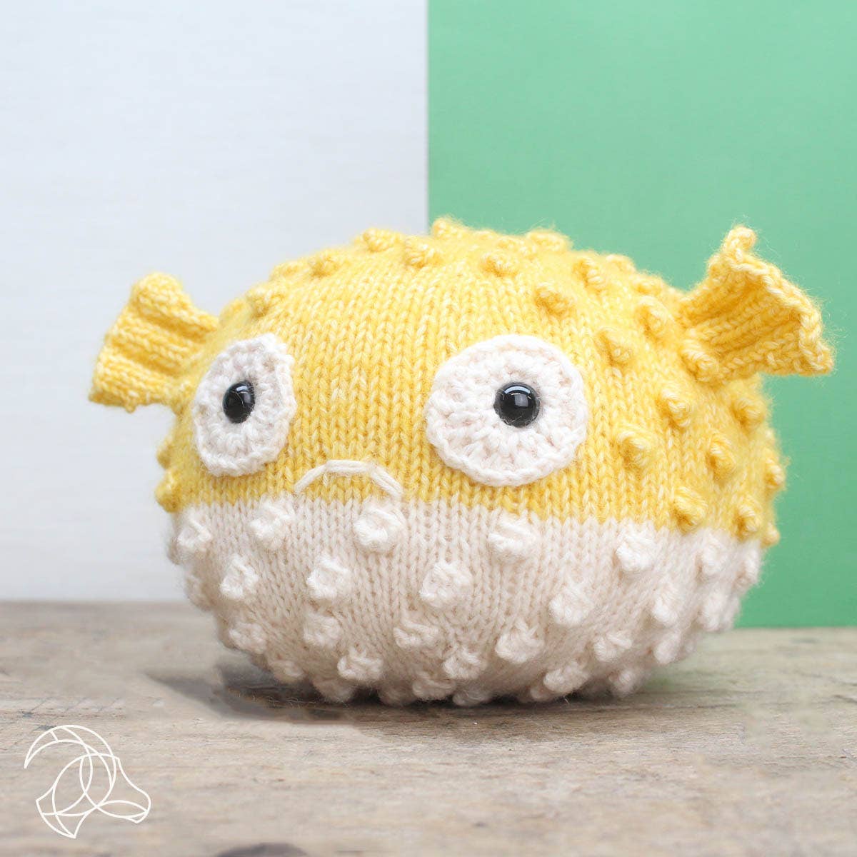 Knitted yellow and white puffer fish toy on a wooden surface with a green and white background