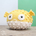 Knitted yellow and white puffer fish toy on a wooden surface with a green and white background