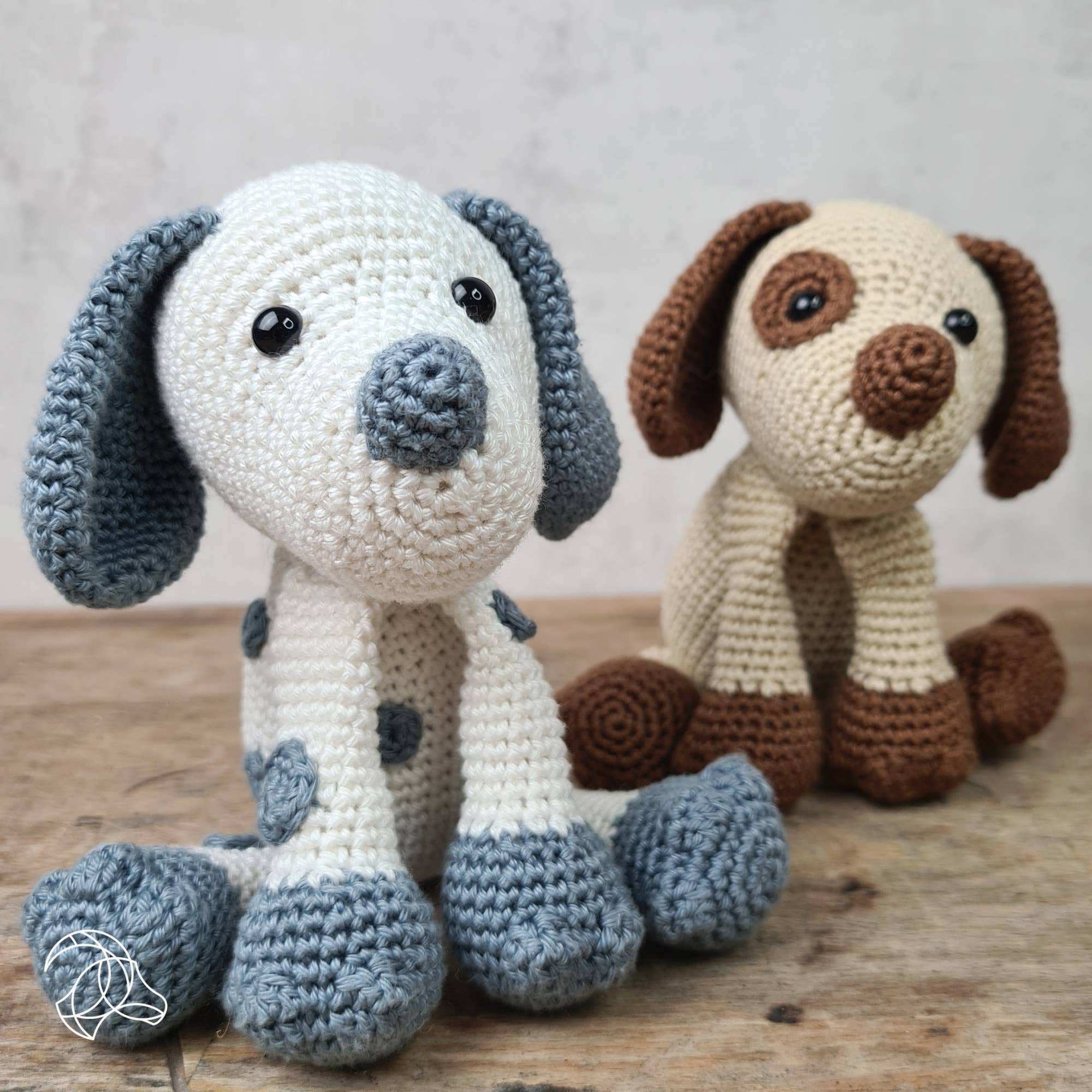 Two crochet dogs, one blue and white with black spots, the other brown and white, on a wooden surface.