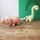 Two crochet dinosaurs, one pink and one green with blue spots, on a wooden surface with a green and white background.