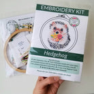 Embroidery kit for hedgehog design held by a hand, with an embroidery hoop and materials in the background.