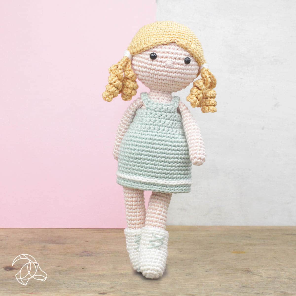 Crocheted doll with blonde hair and a light green dress on a wooden surface with a pink and white background.