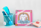 Colorful stationery items including a blue cup with pens, a pink notepad with a cat unicorn design, and a pink clip on a white background.